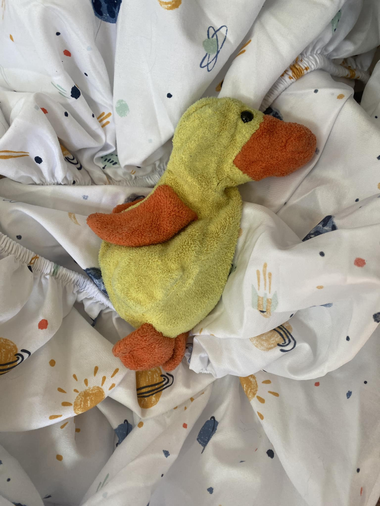 The image is a small yellow duck with orange beak, wings and feet atop a white fitted sheet with stars, planets and suns. 